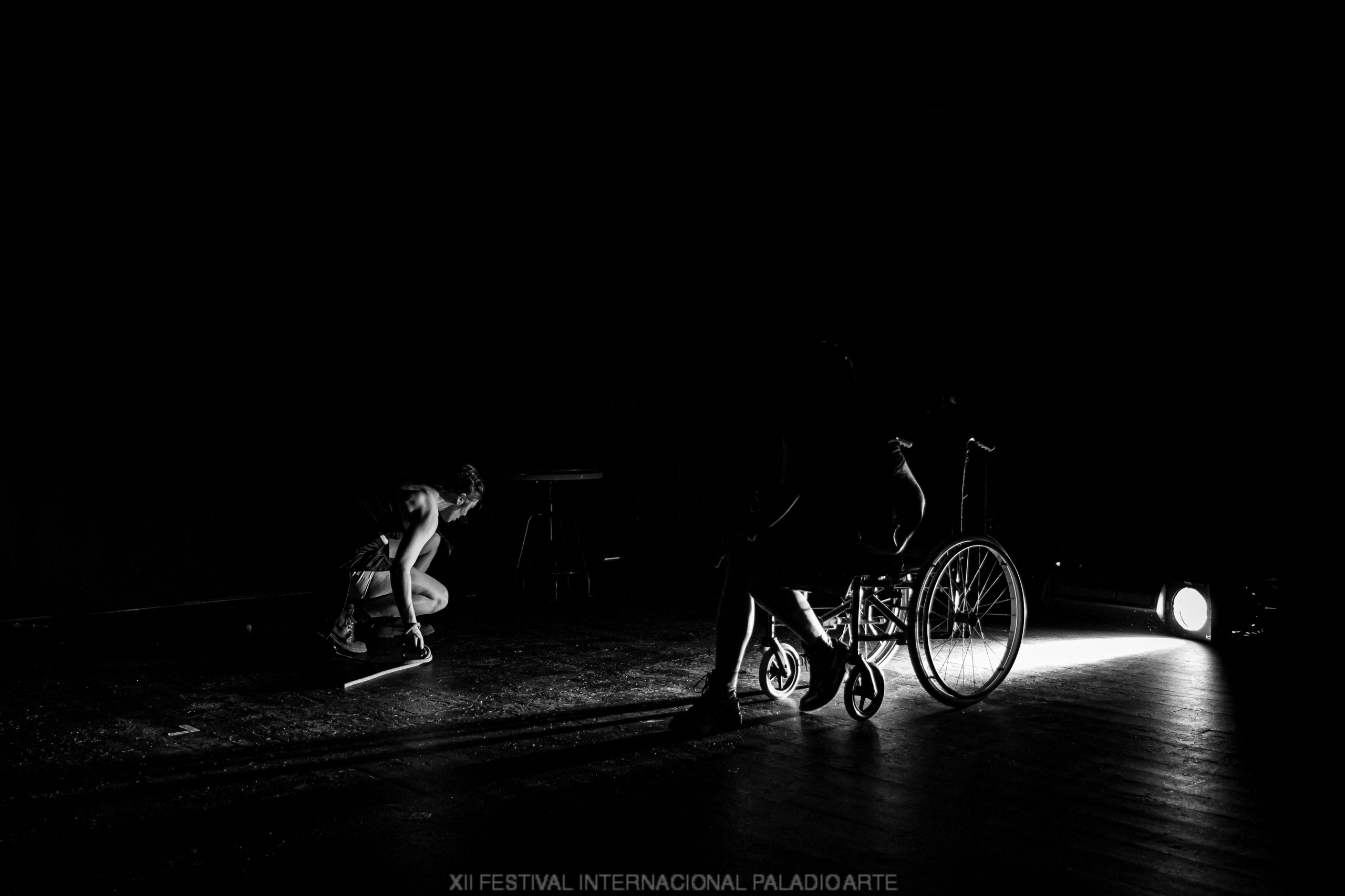 Una silla de ruedas, una persona agachada iluminadas por una sola luz