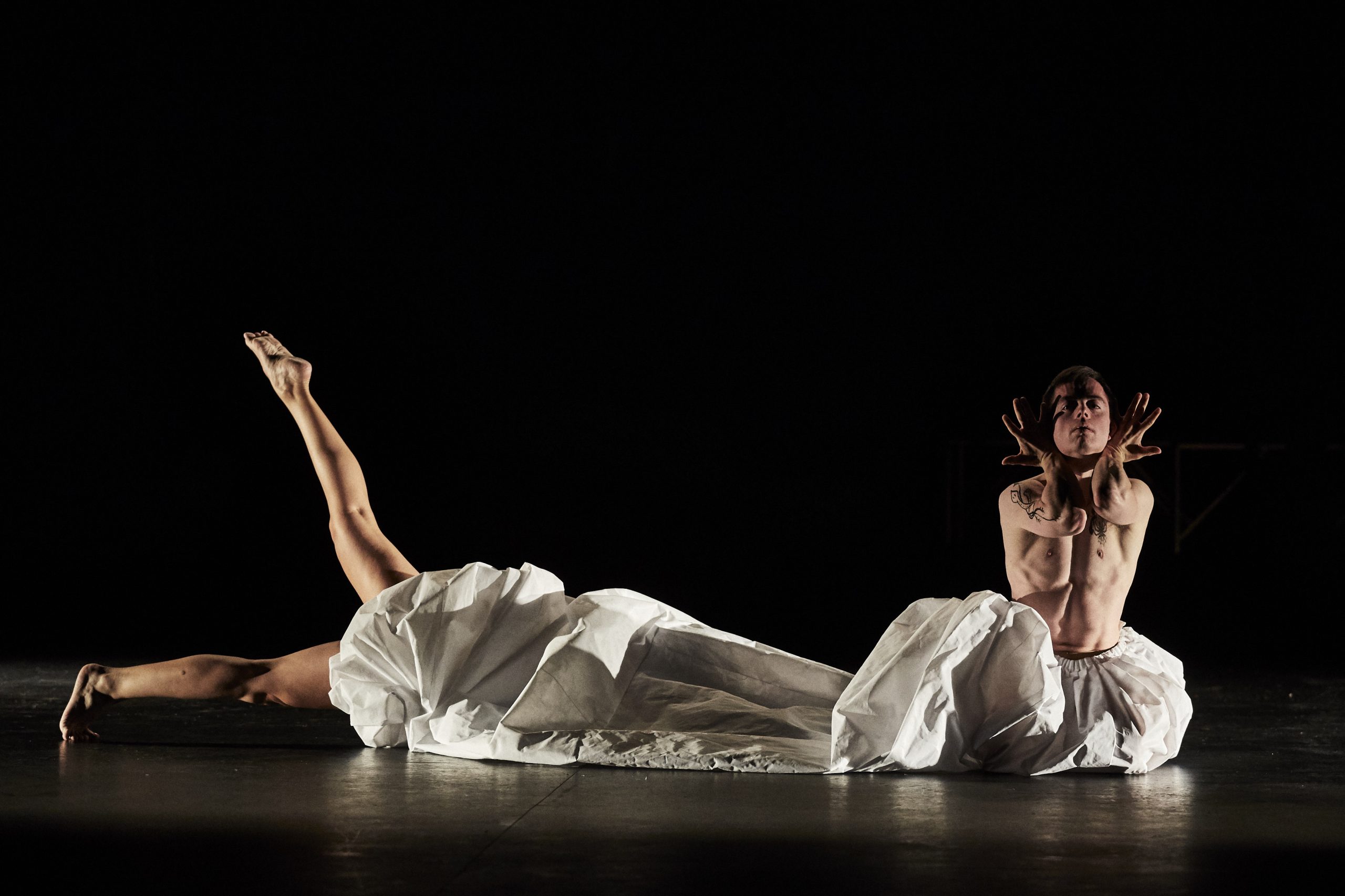 En el escenario dos bailarines y un edredón blanco que tapa el cuerpo de un bailarín tumbado y solo se le ven las piernas
