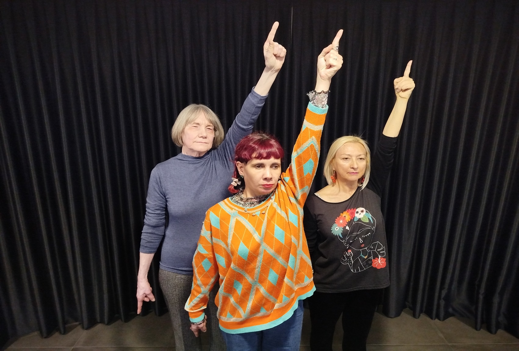 Tres mujeres con jerséis de colores diferentes mirando al frente y con el brazo izquierdo subido y con un dedo señalando al cielo. De fondo un telón negro.
