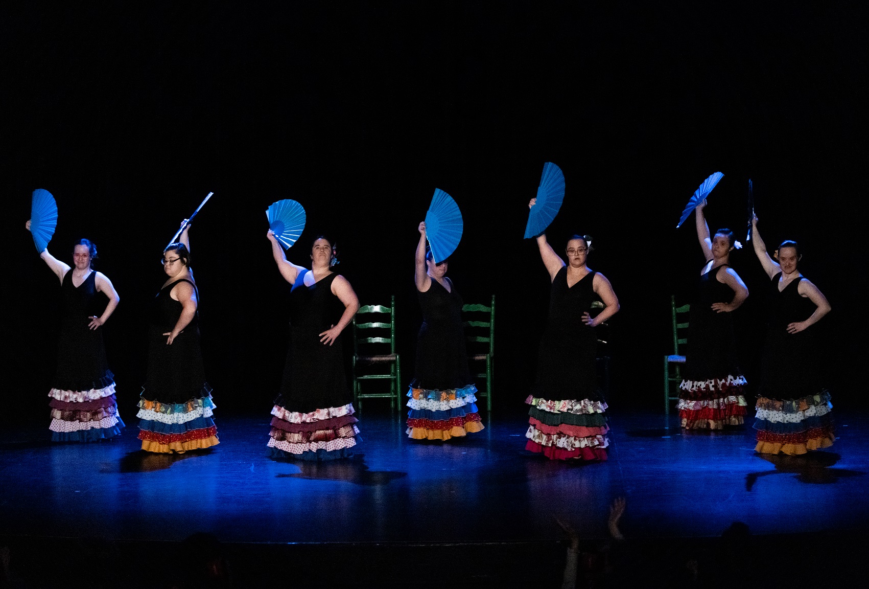 Foto de escena. Un grupo de siete bailaoras tienen un abanico azul en su mano, al fondo sillas de tablao flamenco