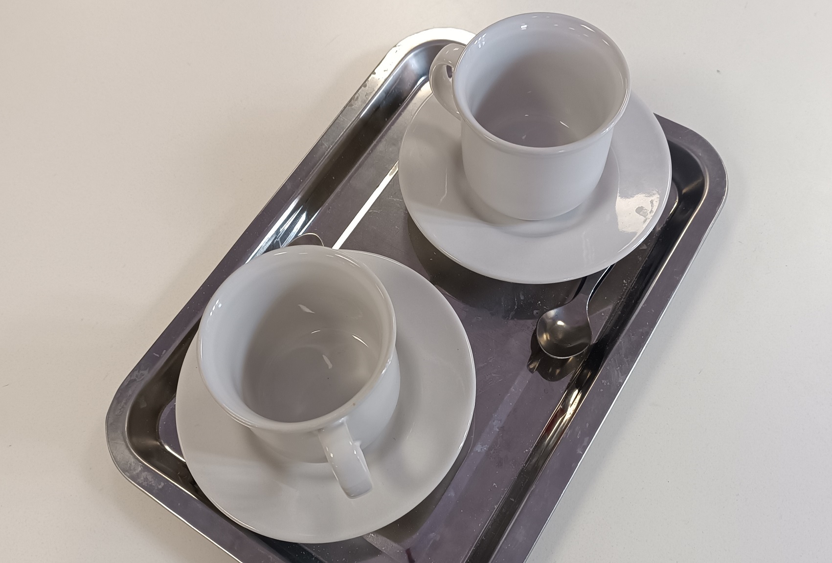 Foto de escena. Dos tazas blancas con sus platos en una bandeja de acero.