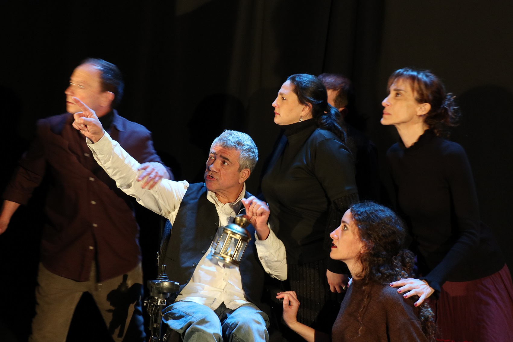 Foto del taller. Un grupo de actores y actrices en el escenario. Miran hacia el lugar que señala un hombre que va en silla de ruedas y tiene en sus manos un farolillo