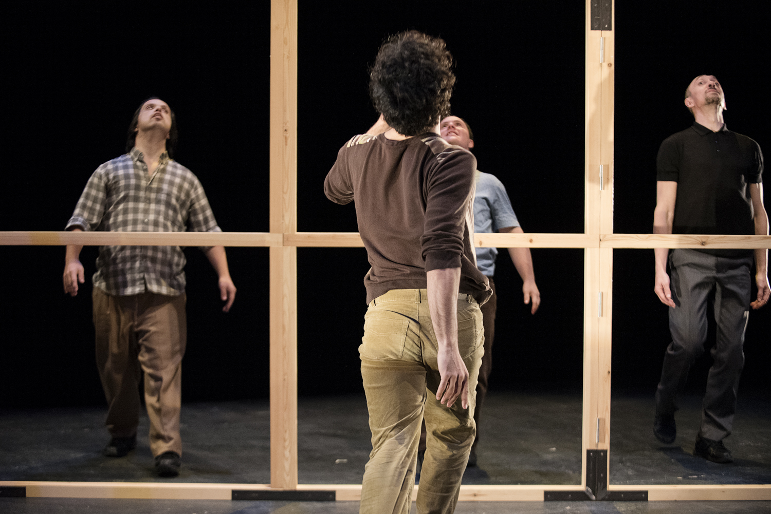 Foto de escena. Cuatro bailarines en escena, vestidos con pantalones y camisas variadas. Tres están de frente a una estructura de madera y la otra está de espalda y mirando a los otros tres.