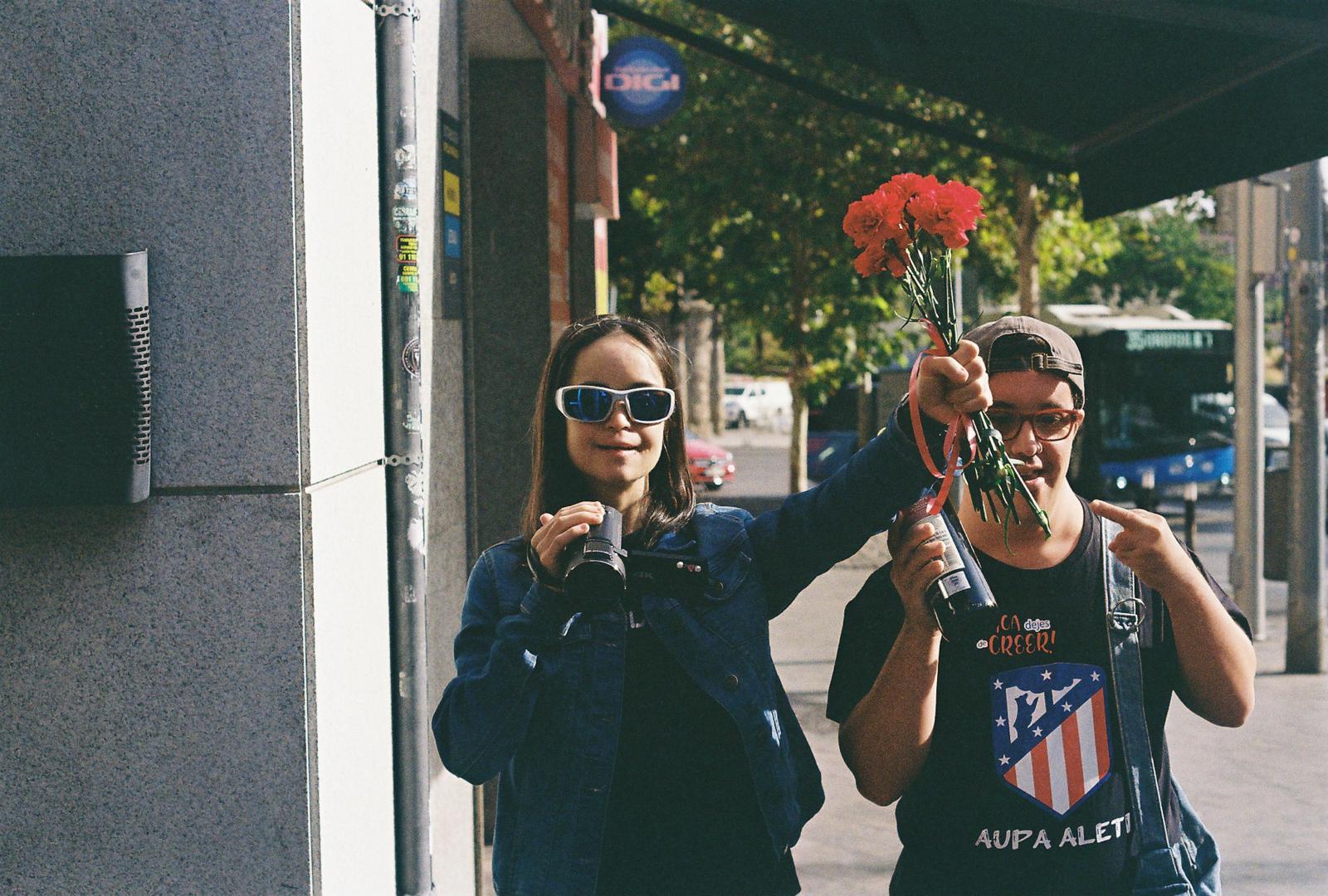 Imagen de cortometraje. Dos personas pasean por la calle. La chica lleva un ramo de claveles rojos y una cámara de video y el chico lleva una botella de vino y hace un gesto con el dedo medio de su mano.