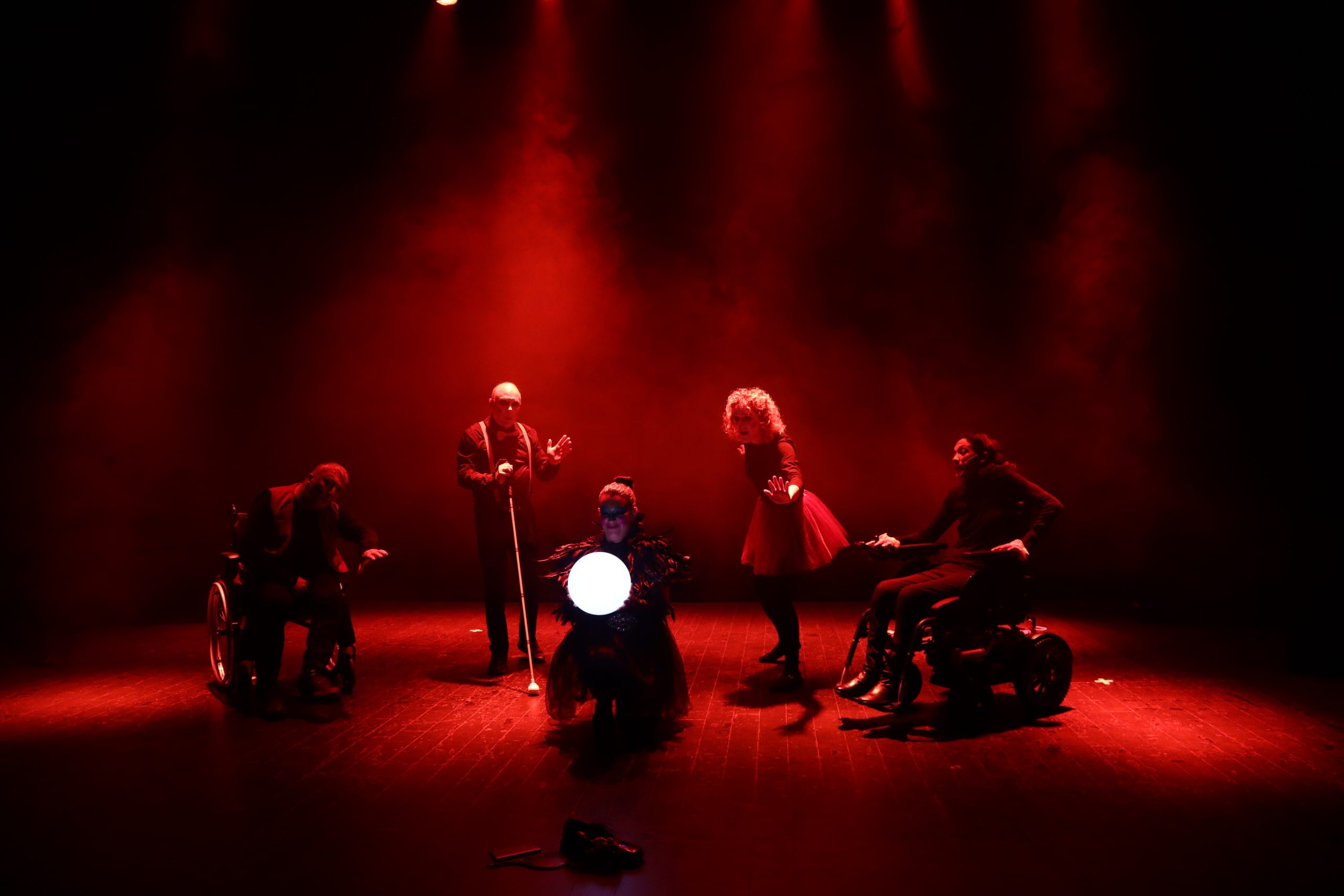 Foto de escena. Cinco personas en escena, la persona en el centro tiene una bola de luz y las otras personas, dos en sillas de ruedas, otra con un bastón guia y una mujer con falda, la rodean y la miran con una luz roja que tiñe todo el escenario