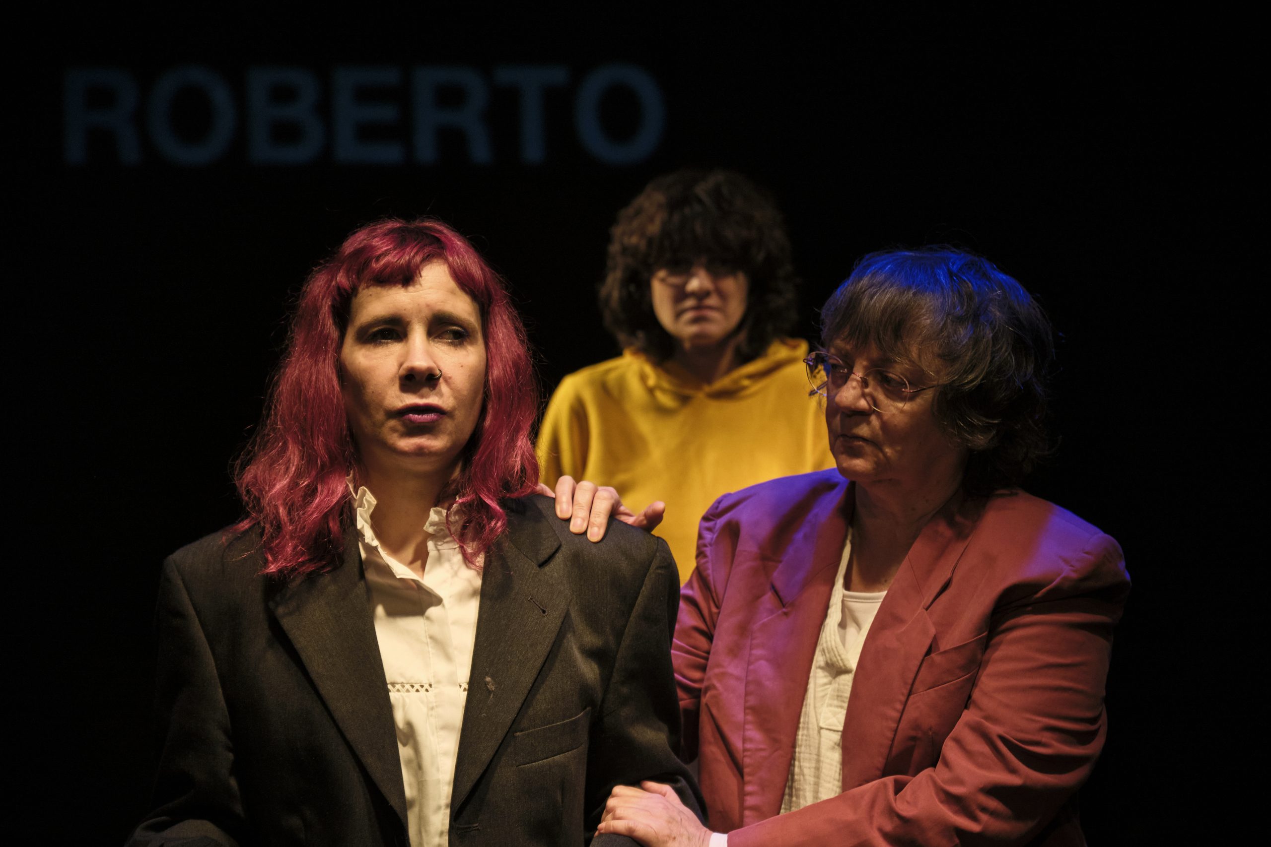Foto de escena. Tres mujeres, dos delante y una atrás en el centro. Una de delante le agarra en brazo a la otra y la de atrás les mira.