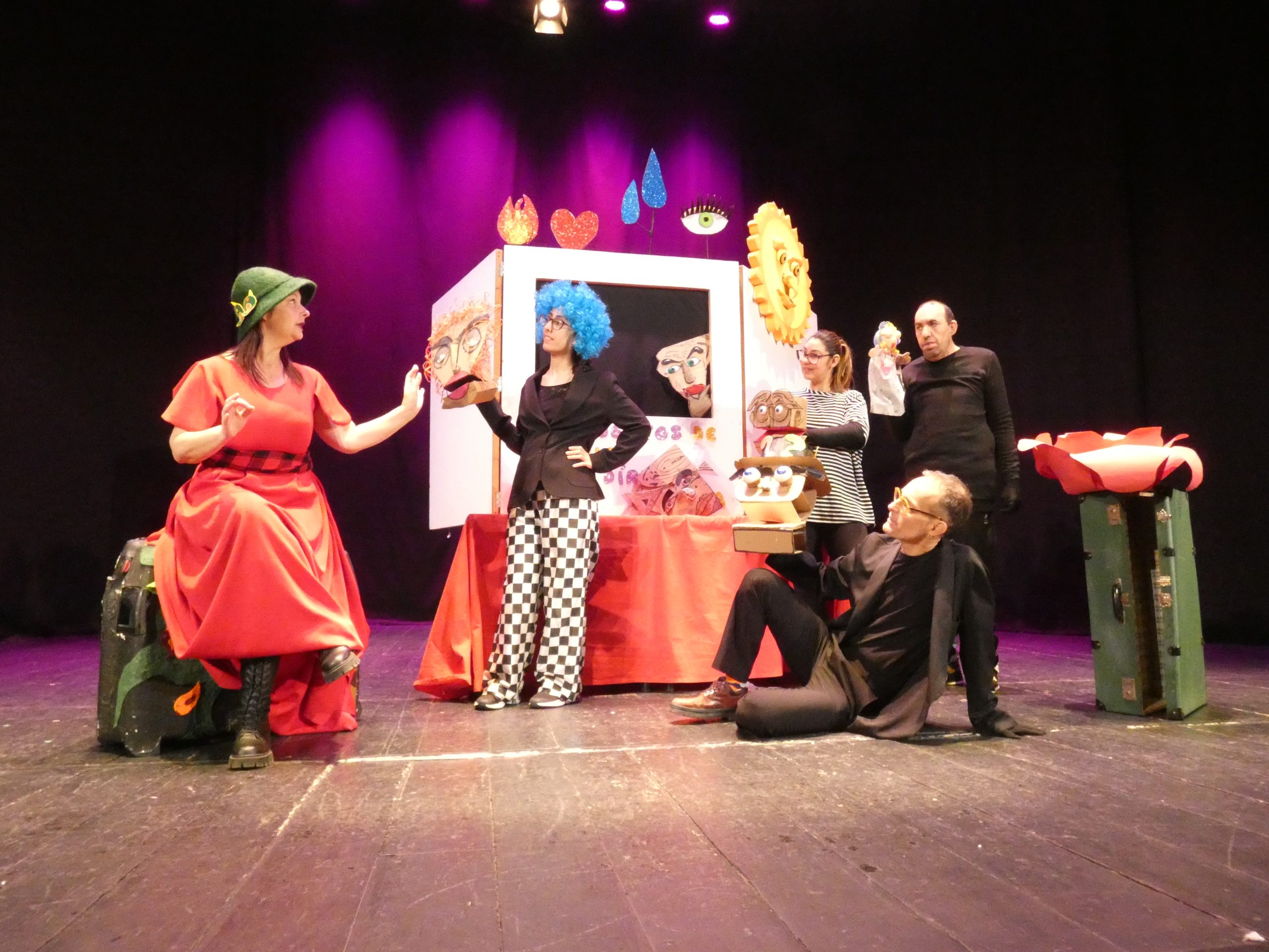 Foto de escena. Un grupo de artistas en el escenario. Al fondo un pequeño teatro del que aparecen unas máscaras.