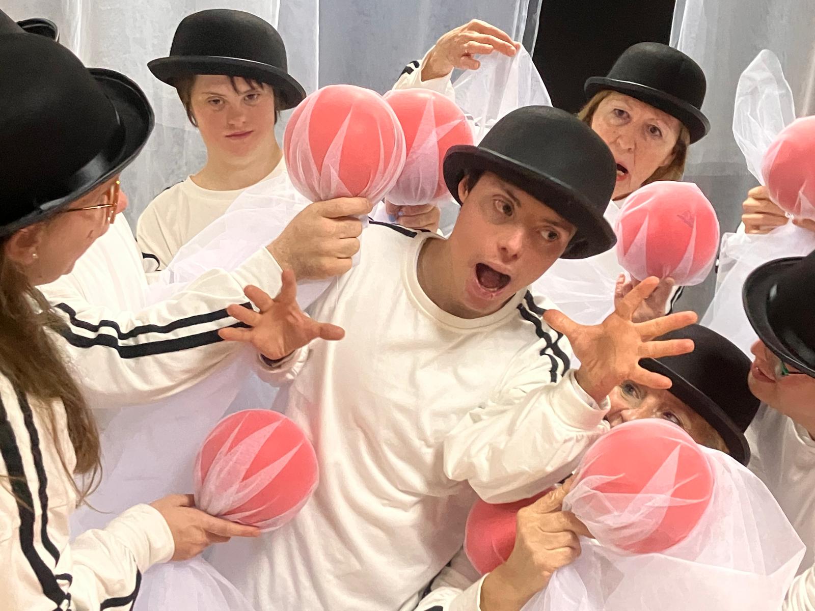 Foto del espectáculo. Un grupo de personas, vestidas con chándal blanco con tiras negras en los laterales, llevan un bombín en la cabeza y una bola roja envuelta en tul