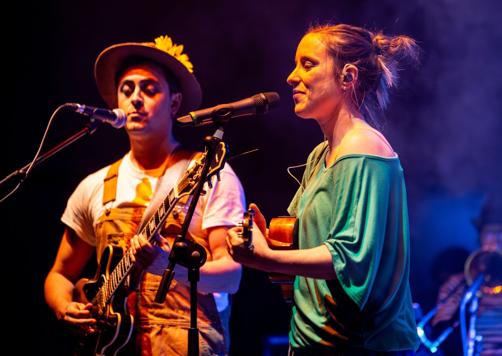 Foto de escena. Un hombre y una mujer cantan. El hombre con peto naranja, un gorro de paja y una guitarra. La mujer con vestido verde y una guitarra.