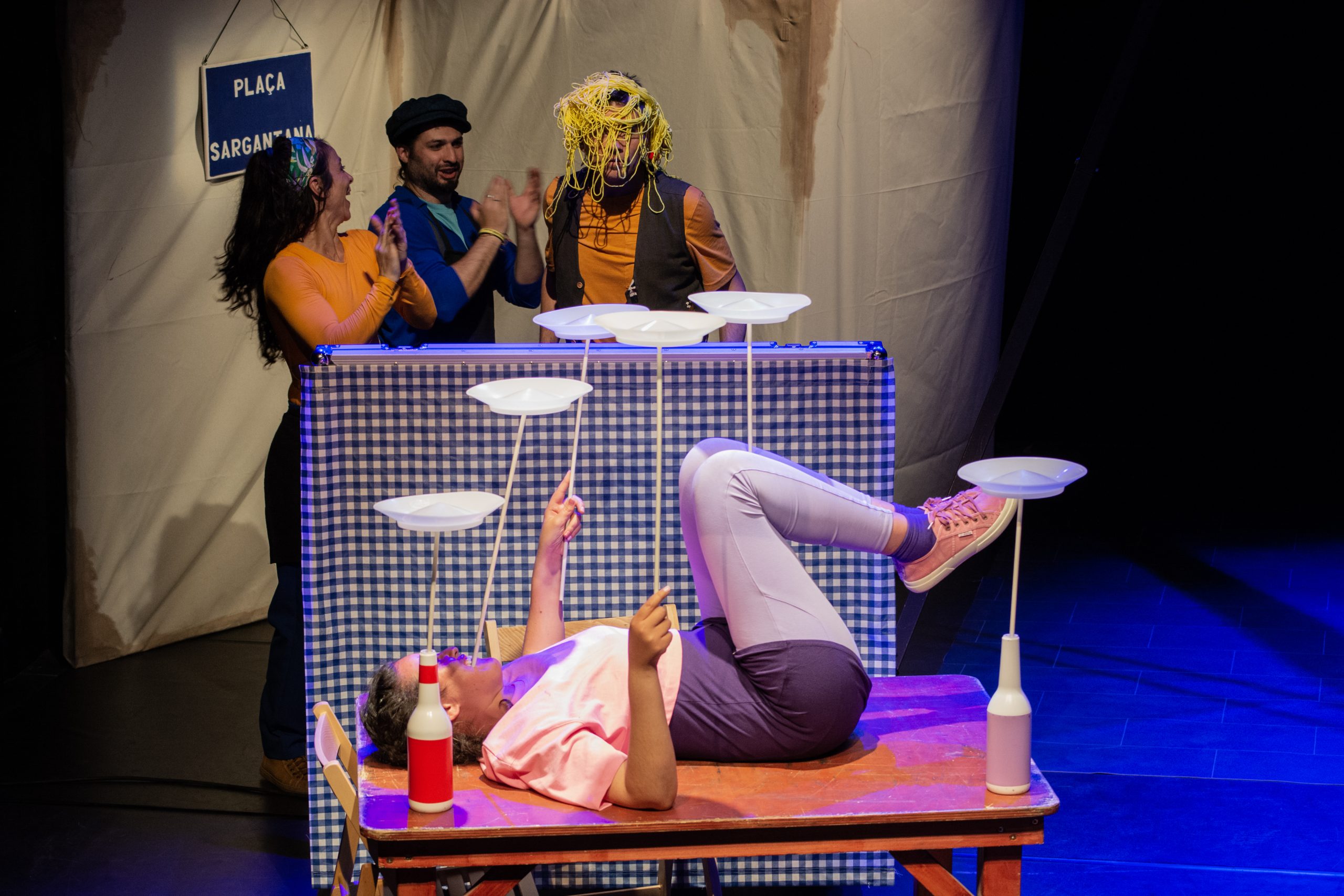 Foto de escena. Encima de una mesa se ve a una persona tumbada con varillas con platos en la boca, en las manos, en las rodillas. Al fondo tres personas miran, una de ellas tiene spaguetti en la cabeza