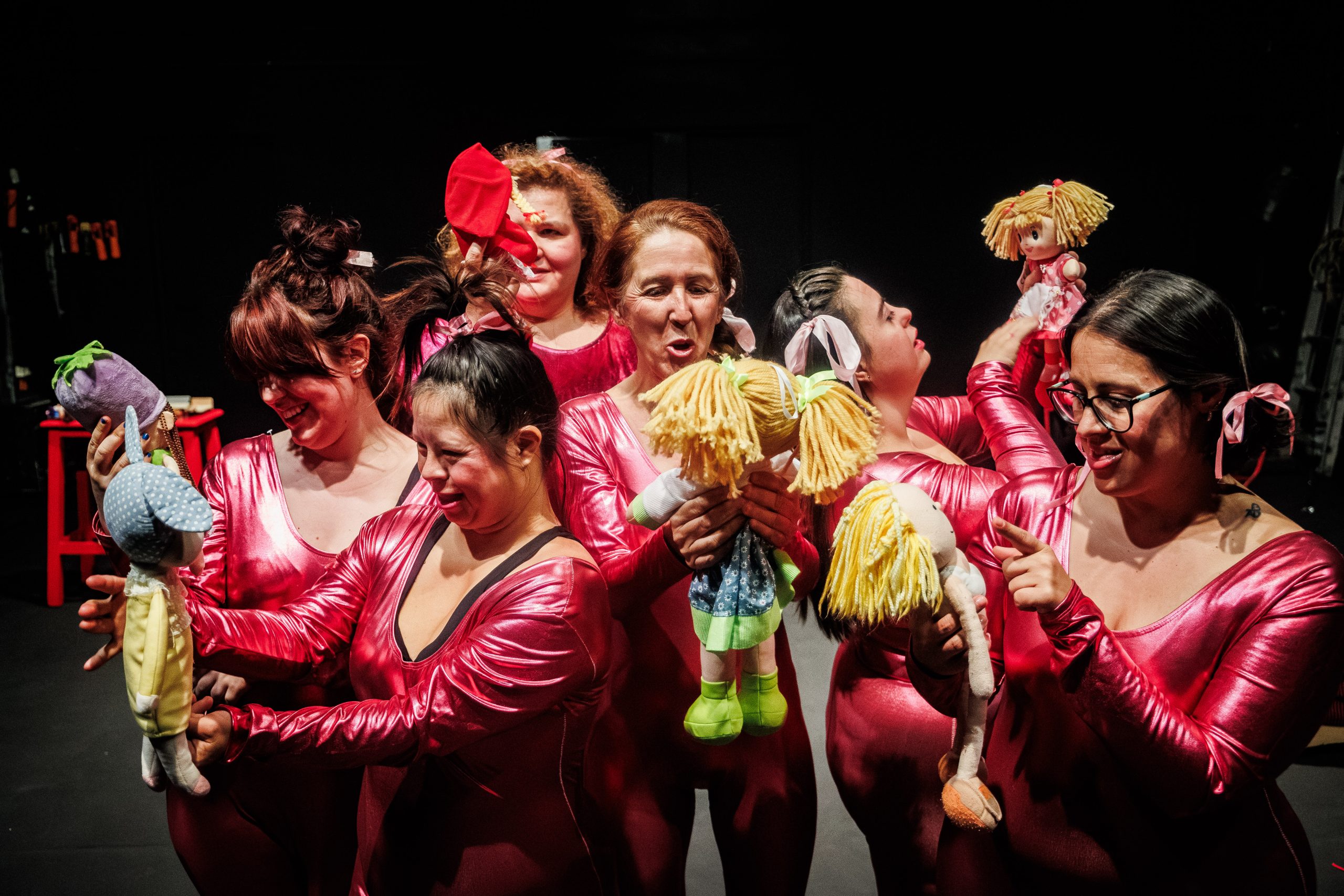 Foto de escena. Un grupo de mujeres, vestidas con unos trajes rosas brillantes, tienen en sus manos muñecas y unas hablan con otras.