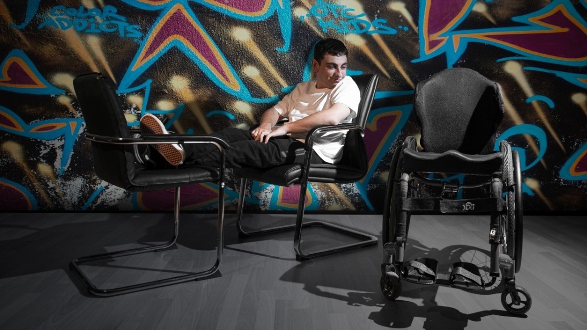 Imagen del espectáculo. Un chico sentado en una silla con los pies en otra silla mira una silla de ruedas vacía.