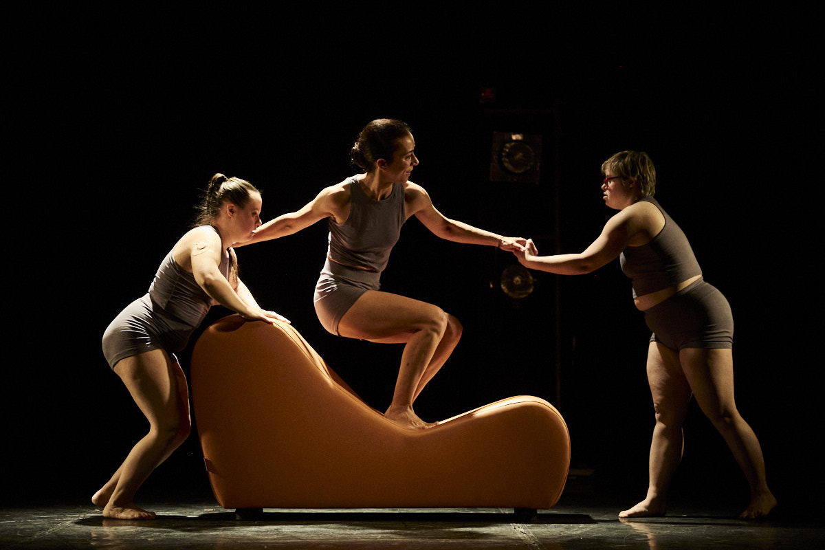Foto de escena. Tres bailarinas, una encima de un sofá moderno, y las otras a la izquierda y derecha de ella.