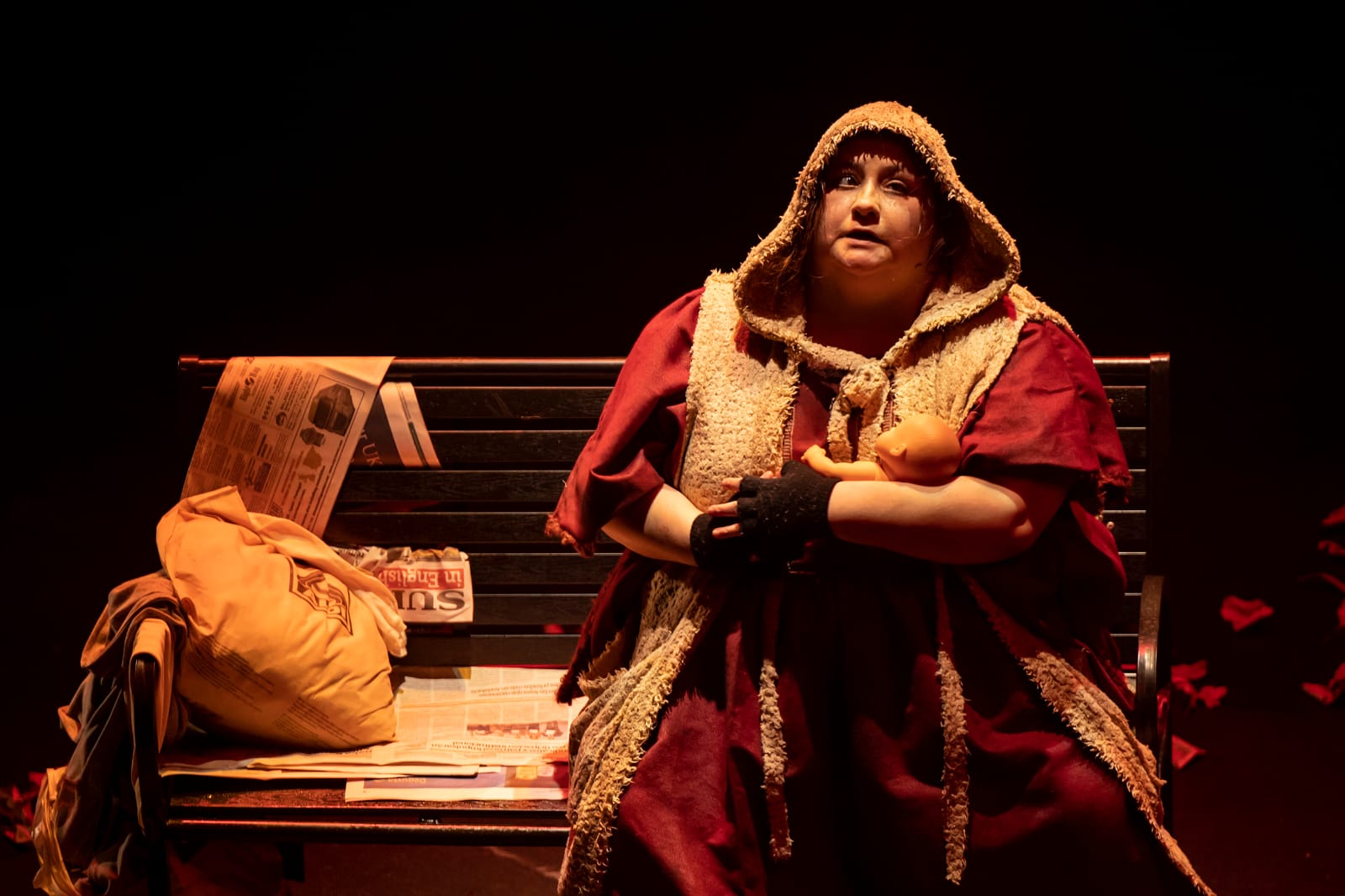 Imagen del espectáculo. Una mujer vestida de rojo y con una capucha que cubre su cabeza está sentada en un banco de parque. Al lado una bolsa con sus cosas personales.